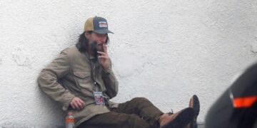 Киану Ривс како бездомник со седа брада и долга коса пуши цигара пред клуб